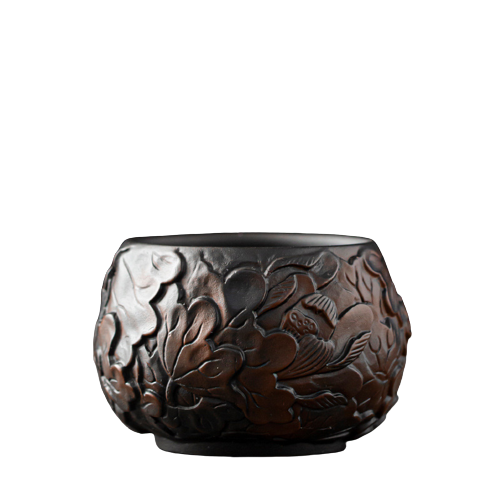 A black ceramic teapot with a floral pattern, resembling a lotus flower, on a plain background.