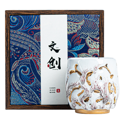 A white ceramic coffee cup with gold and red floral patterns, accompanied by a gift box with a traditional design.