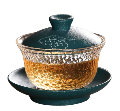A clear and green colored crystal Gaiwan with a lid and a handle, filled with a golden-brown liquid, presumably tea.