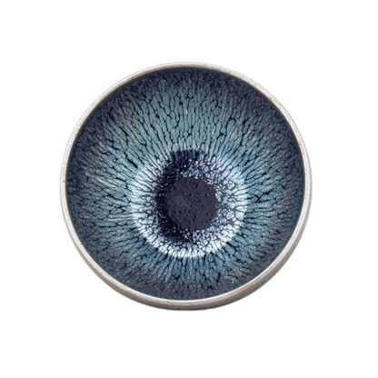 A circular ceramic teacup with a blue and black speckled pattern in the center, resembling an eye, with a metallic rim.